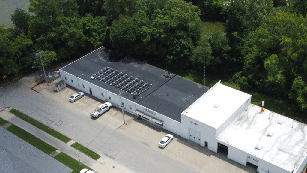 Drone image of Keusch Glass facility highlighting a flat-roof commercial solar array surrounded by green trees and neighboring industrial buildings, emphasizing renewable energy adoption.